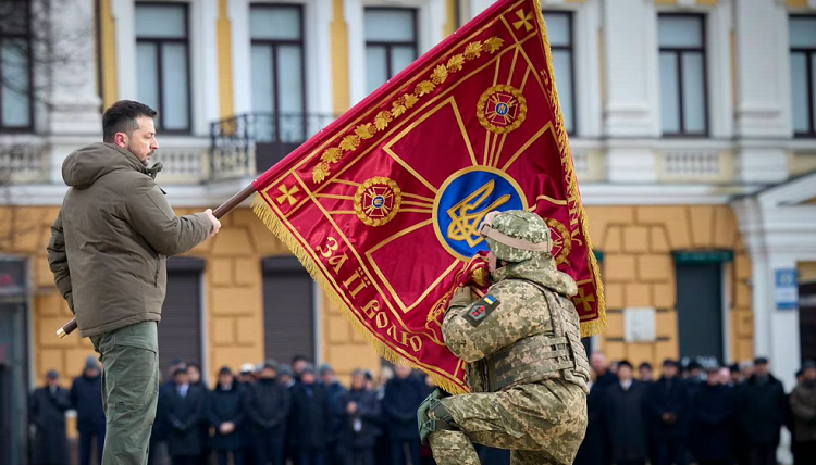 الرئيس الأوكراني فولوديمير زيلينسكي يحمل علم وحدة عسكرية بينما يقبلها ضابط في كييف بأوكرانيا الشهر الماضي